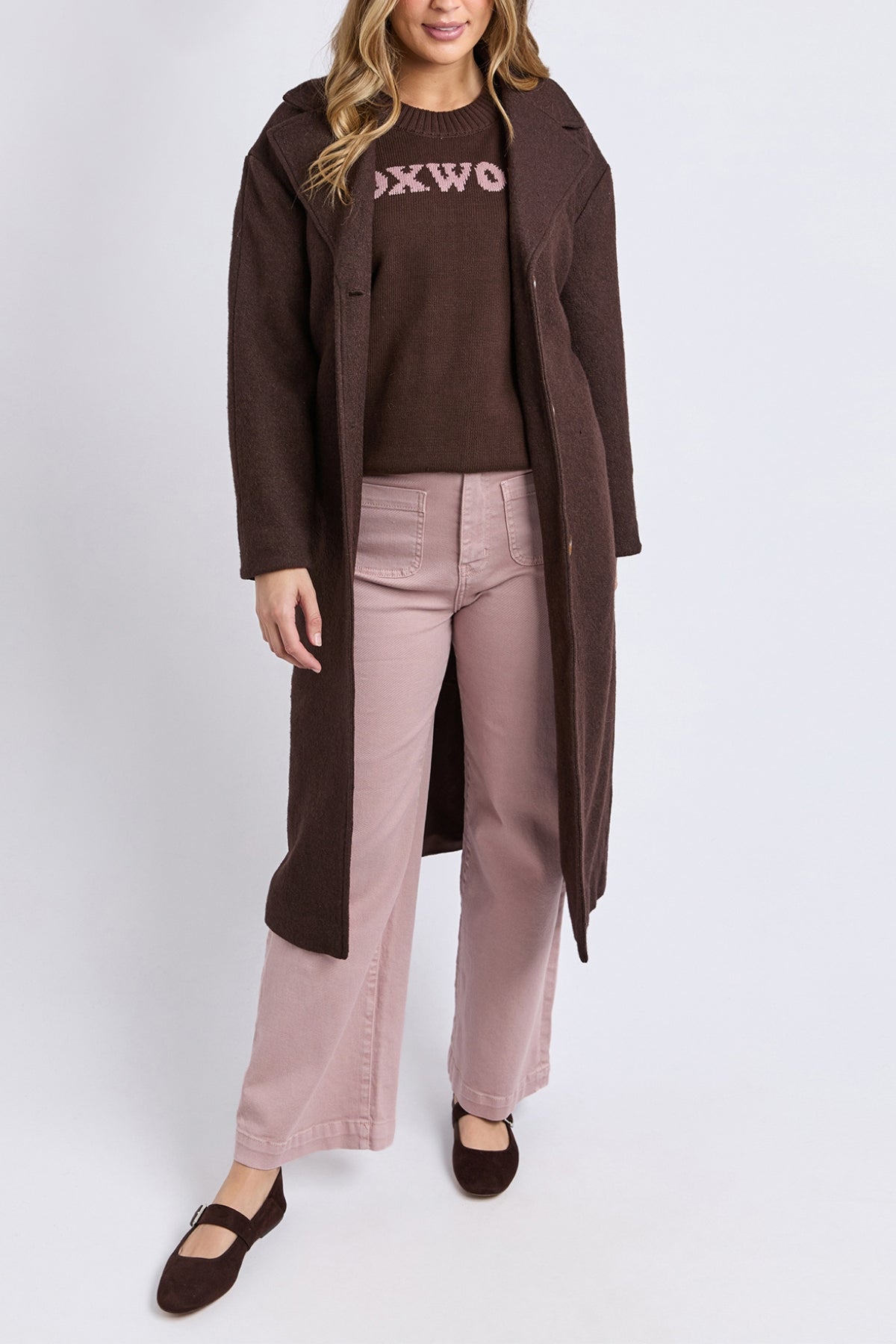 A person in a Foxwood Whistler Coat (wool-polyester blend), a brown sweater with "OXWOR" in pink, light pink wide-leg pants, and brown flats stands against a white background—a chic twist on women’s outerwear.