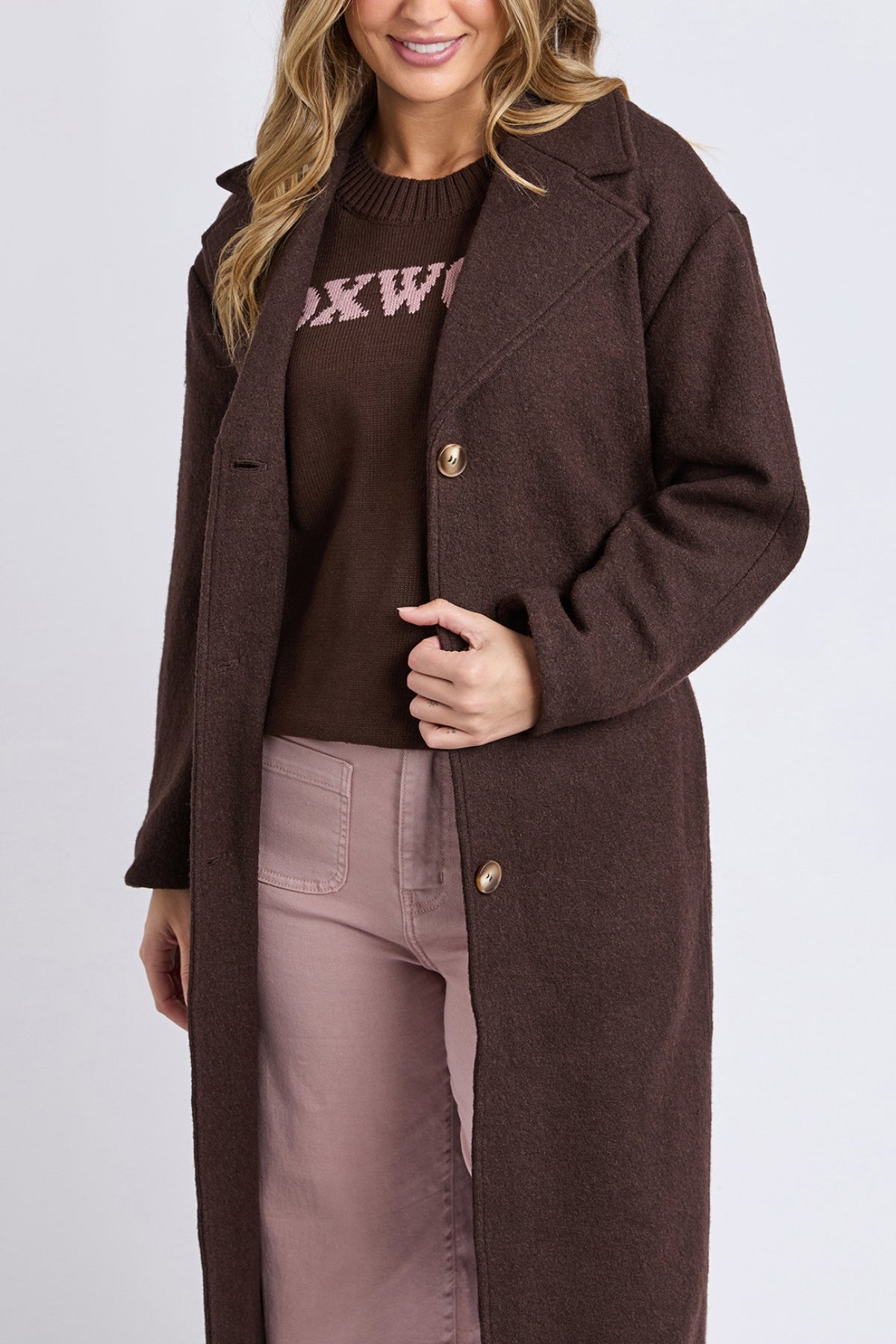 A woman with wavy blonde hair, smiling softly, wears the Foxwood Whistler Coat—made from a wool-polyester blend—over a brown sweater with pink writing and light pink pants. Only her upper body is visible for a chic outerwear look.