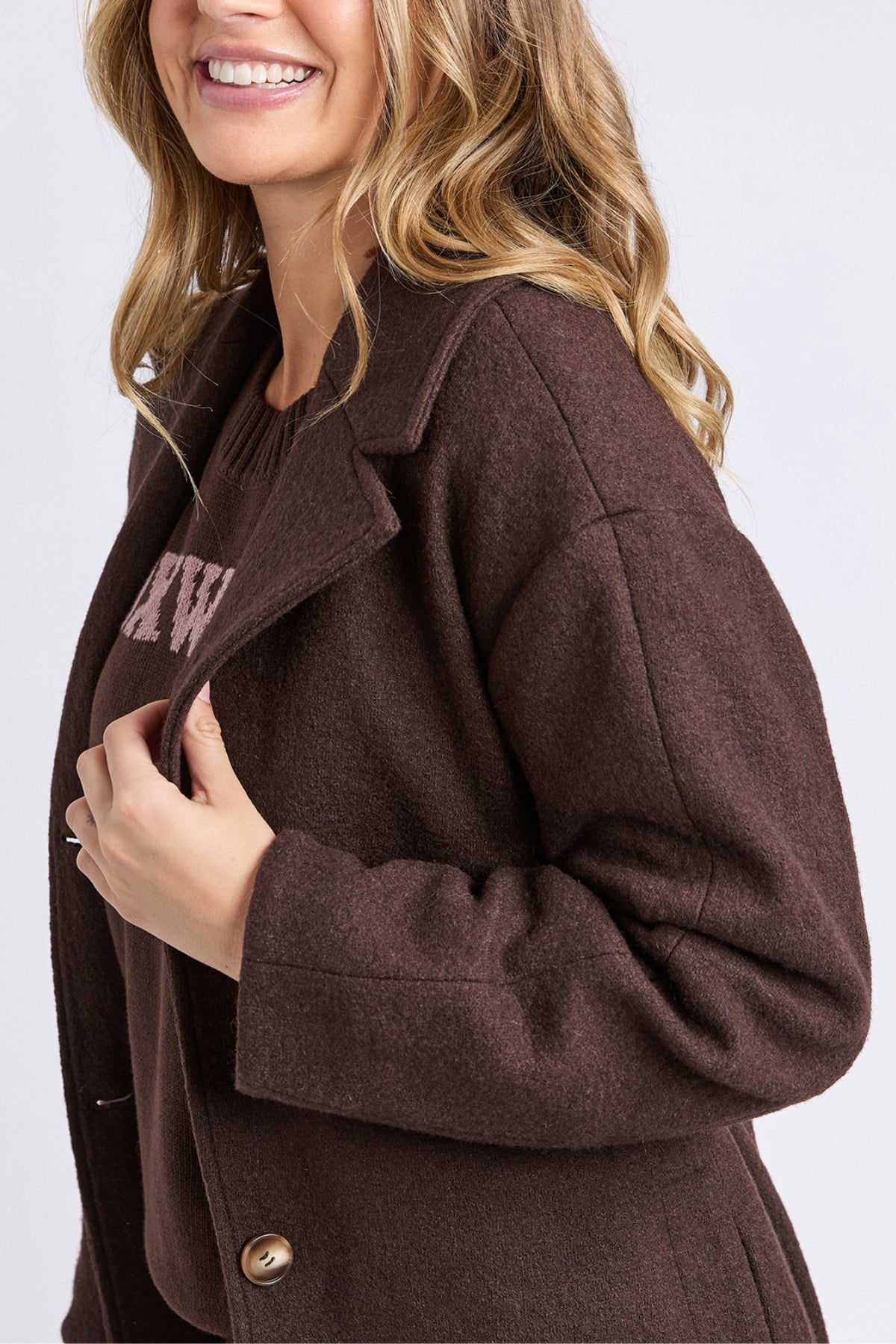 A woman with long, wavy blonde hair smiles while wearing the Foxwood Whistler Coat, a cozy women’s wool-polyester blend outerwear. She turns slightly, showing off the coat’s lapel and buttons against a light background.