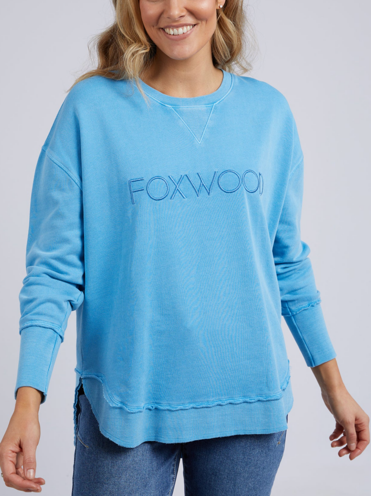 A woman smiles, wearing the Foxwood Simplified Crew—a loose-fitting, light blue crew neck jumper with an embroidered Foxwood logo—paired with blue jeans. She stands against a plain background with her hands relaxed at her sides.