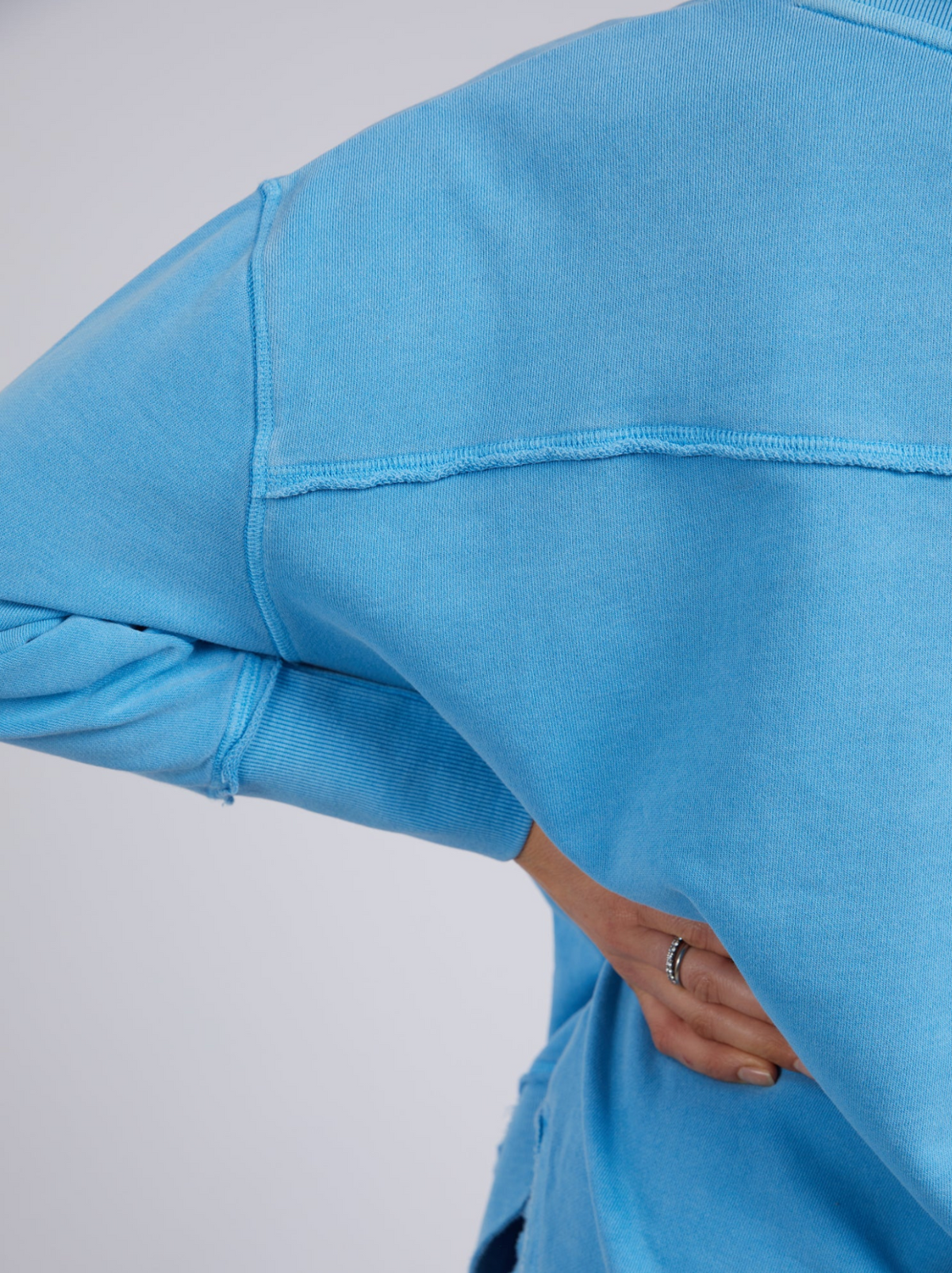 Model wears the Foxwood Simplified Crew, a light blue jumper with visible seam details and an embroidered Foxwood logo. Upper body and part of arm are shown, with one hand on hip.