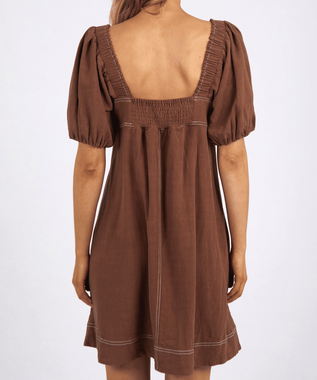 Against a plain backdrop, someone wears the Foxwood Florence Dress with its brown, puff-sleeves, square neckline, and back smocked details. The knee-length dress provides a lightweight comfort as their hair is tied back.