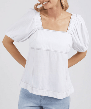 A woman smiles with her hands behind her back, wearing the Foxwood Florence Top—white with a square neckline, short puff sleeves, and contrast stitching—paired with light blue jeans against a plain backdrop.