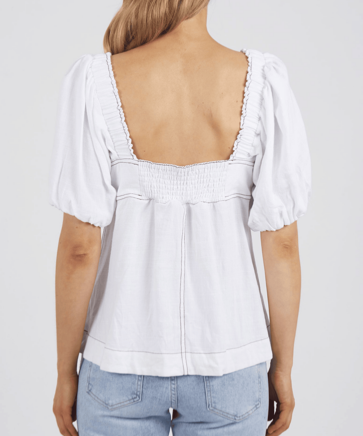 A woman photographed from behind wears the Foxwood Florence Top—a white, short-sleeve blouse with a square neckline, low square back, and puffed sleeves—paired with light blue jeans with contrast stitching.