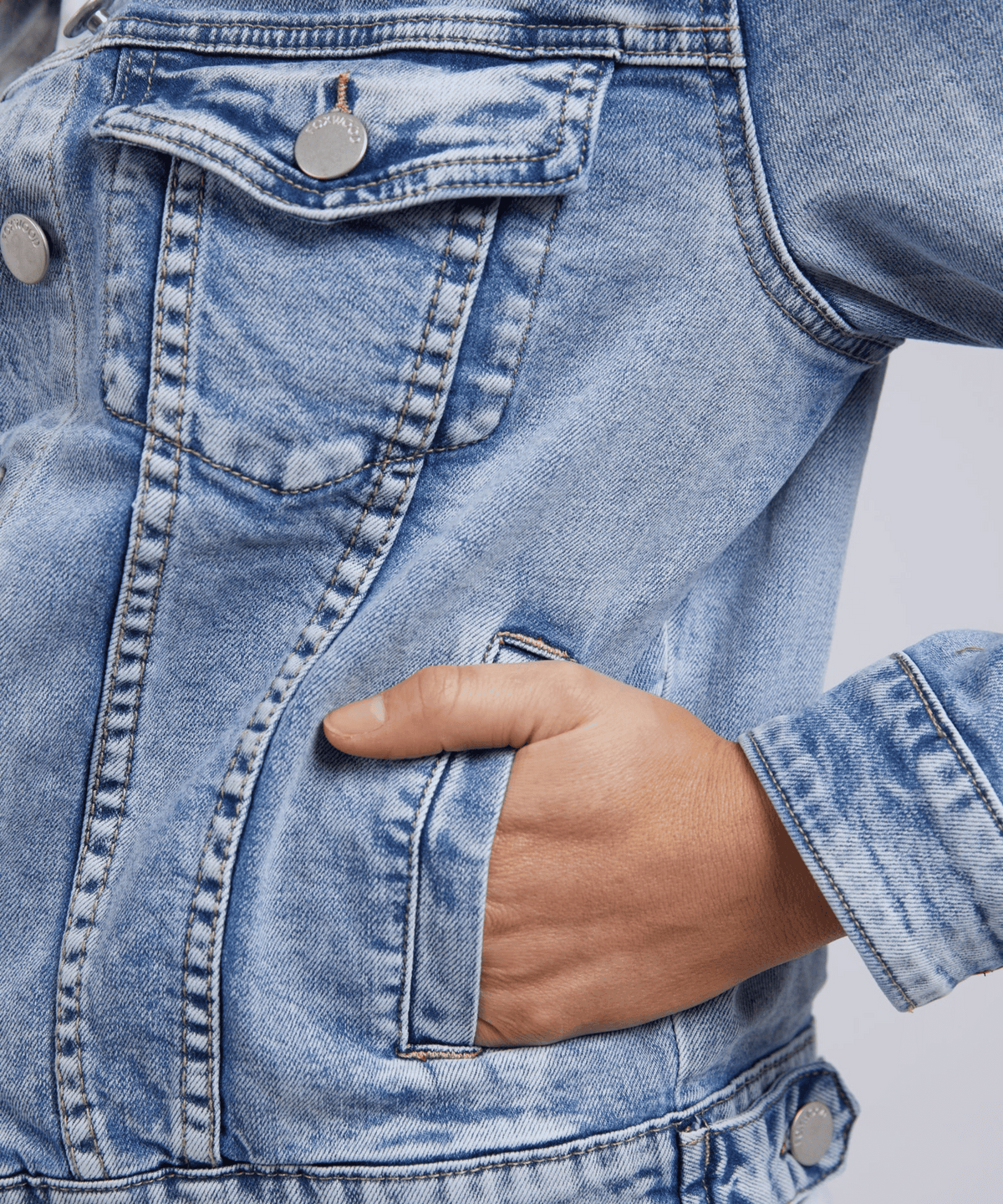 A person wears the Foxwood Georgia Denim Jacket, a smart-casual staple in light blue denim with silver buttons. The cotton-elastane blend has a subtle worn pattern, and their hand rests comfortably in the pocket against a neutral backdrop, emphasizing this versatile piece.