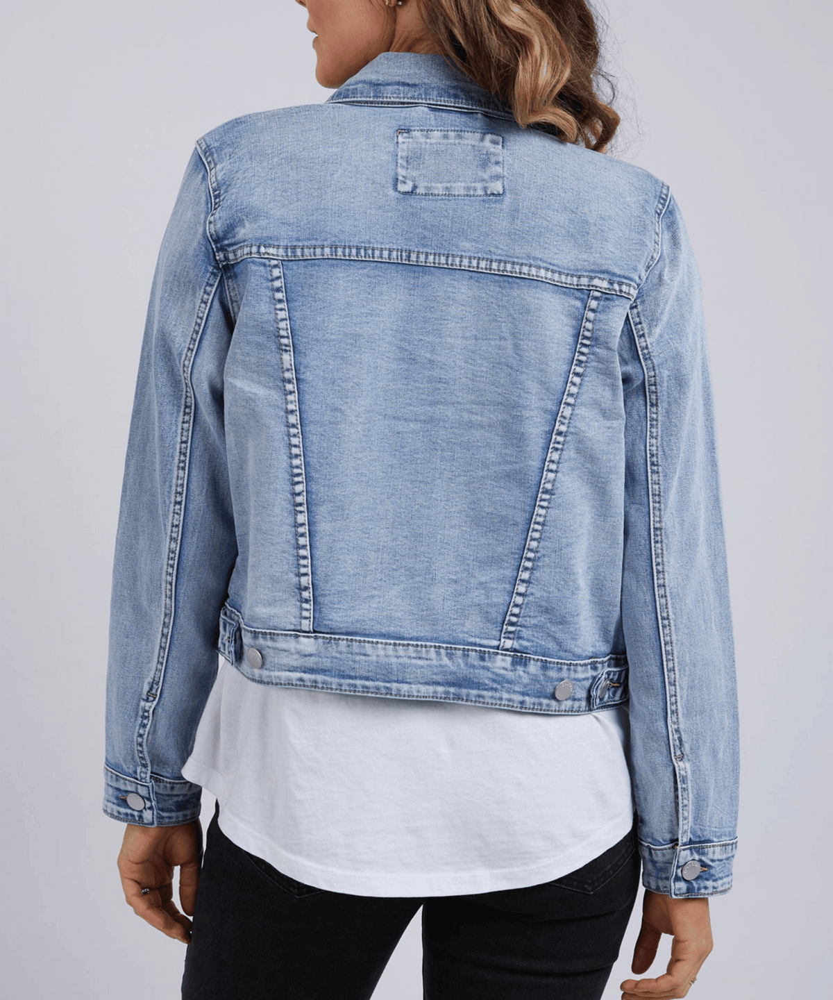 A person with wavy hair is seen from the back, wearing a smart-casual Foxwood Georgia Denim Jacket in light blue cotton elastane, layered over a white shirt and black pants against a plain light gray background.
