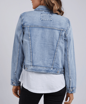 A person with wavy hair is seen from the back, wearing a smart-casual Foxwood Georgia Denim Jacket in light blue cotton elastane, layered over a white shirt and black pants against a plain light gray background.
