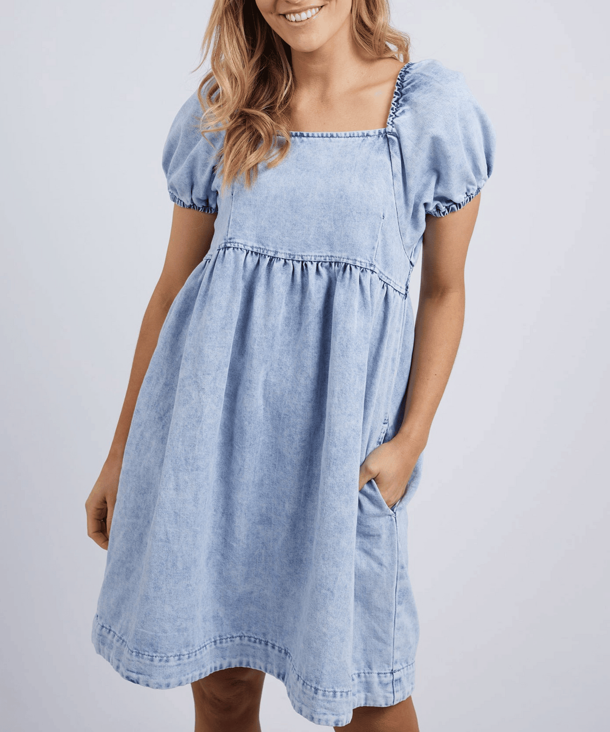 Woman in a light blue Foxwood Jade Textured Dress with short sleeves, square neckline, and puffed sleeves, smiling with one hand in her pocket against a plain light background.