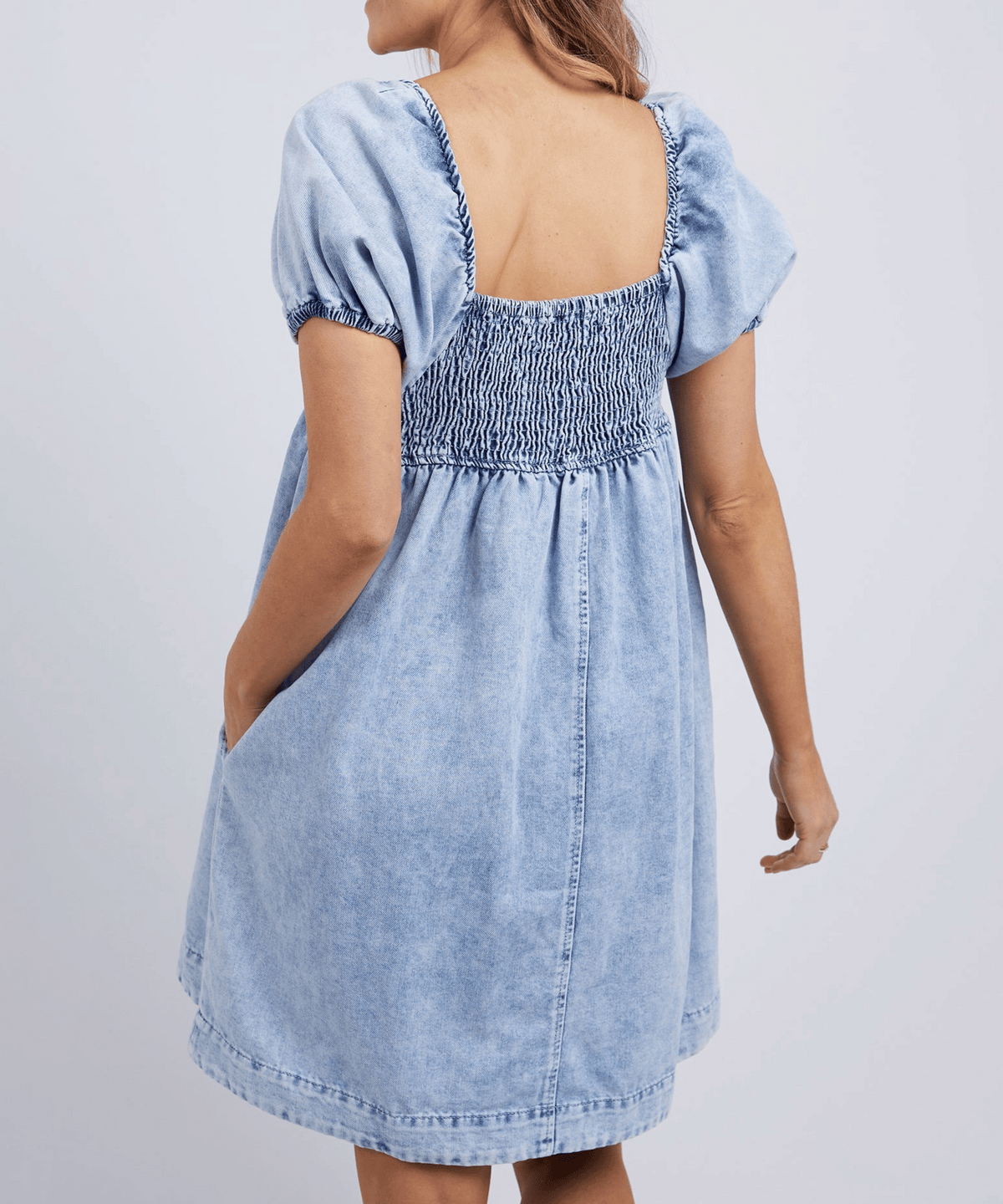 A woman is seen from behind wearing the Foxwood Jade Textured Dress, a light blue short-sleeve cotton dress with elastic smocking on the upper back and sleeves, a square neckline, vintage wash finish, and side pockets.