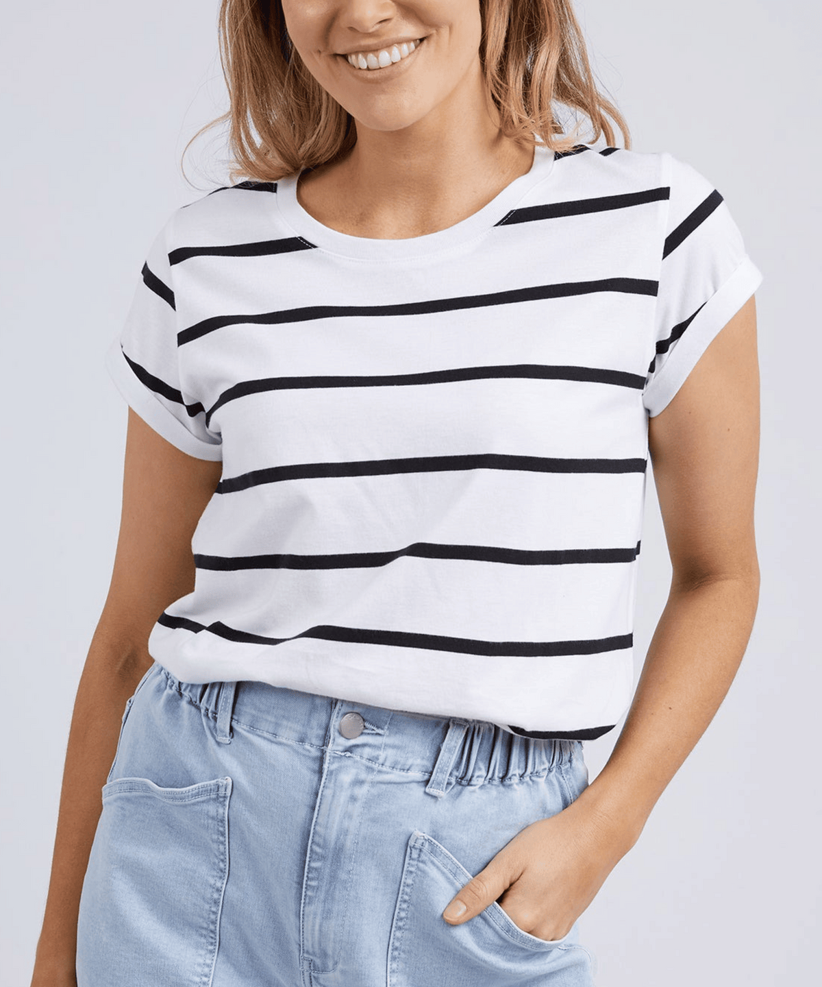 Wearing a Foxwood Manly Stripe Tee made of 100% soft prima cotton, the person pairs it with light blue high-waisted jeans, casually resting one hand in their pocket while smiling warmly against a plain, light-colored backdrop.
