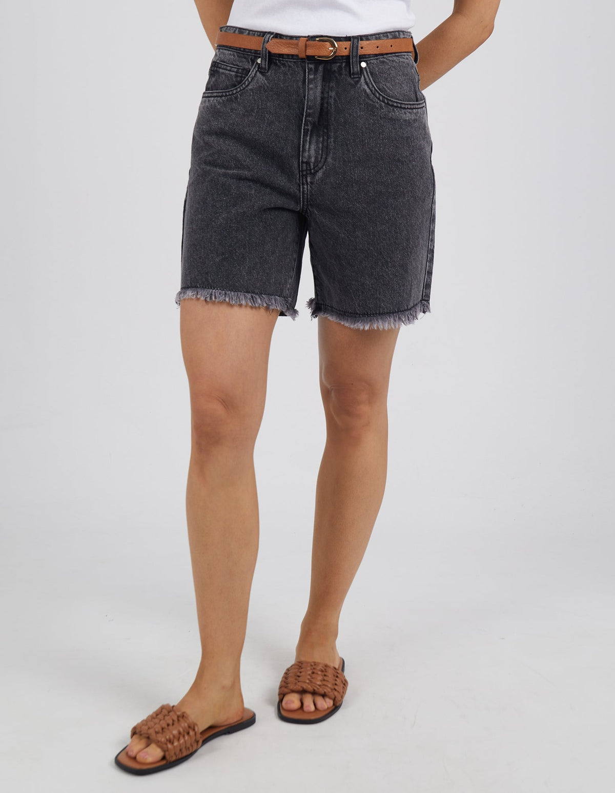 Someone stands against a plain white background wearing Foxwood Millie Short—high-waisted black denim shorts with a frayed hem—paired with a brown belt, white top, and brown woven sandals.