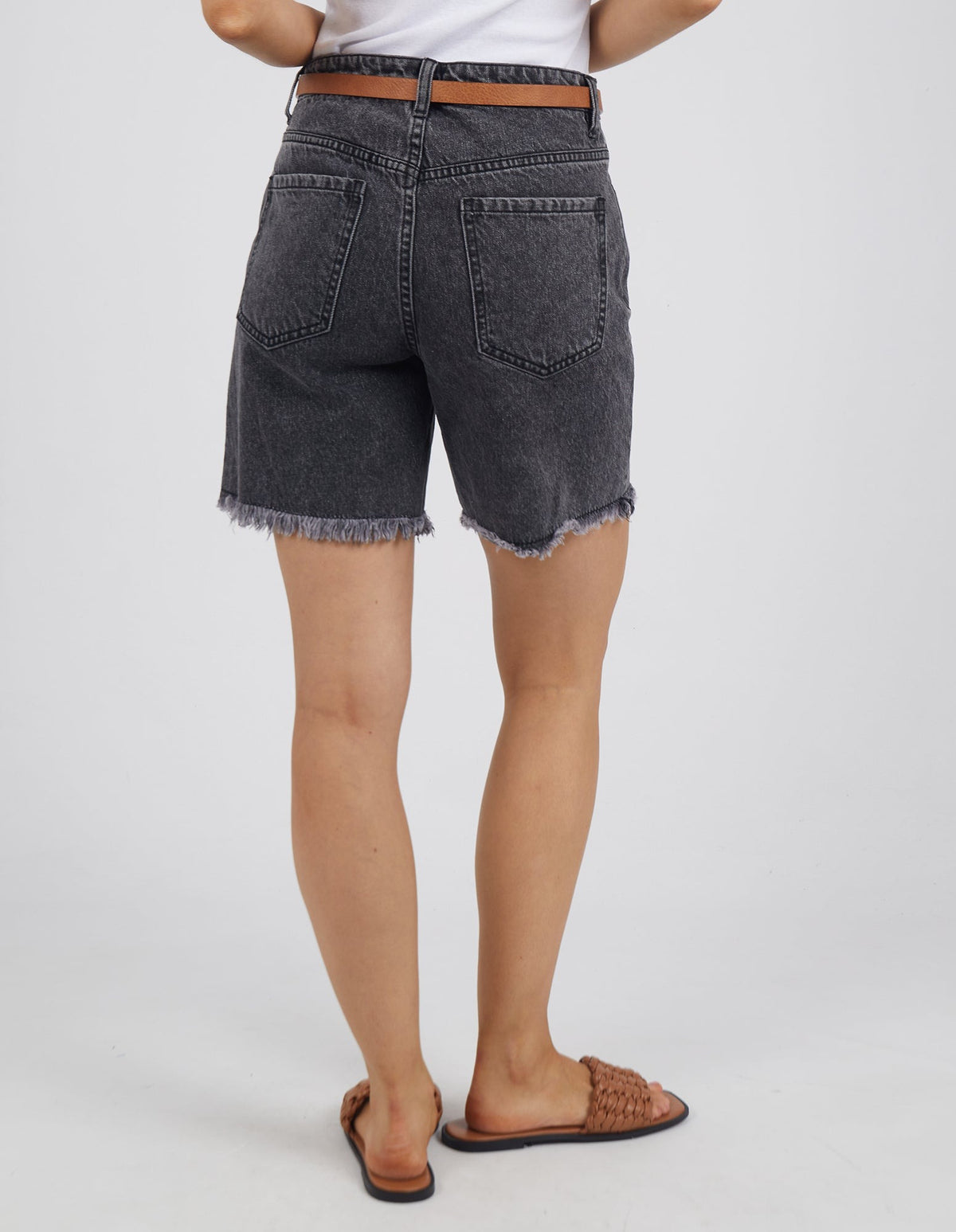 Person standing with their back to the camera, wearing a white shirt and the Foxwood Millie Short—black denim shorts with a frayed hem—paired with a brown belt and brown woven sandals against a plain background.
