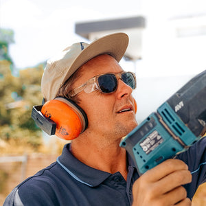 A man wearing SafeStyle Fusions Champagne Frame Tinted Lens safety glasses, orange earmuffs, and a beige cap operates a power tool outdoors, focused on his work with trees and a building blurred in the background.