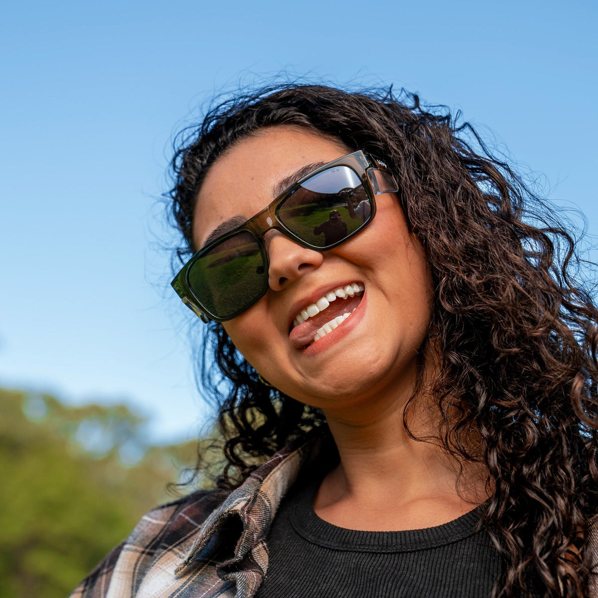 A person with curly hair wears SafeStyle Fusions Green Frame Polarised Lens sunglasses with UV400 protection and sticks their tongue out playfully, basking outdoors under a clear blue sky surrounded by lush green trees.