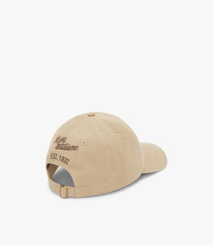 The RM Williams Longhorn Cap is shown from the back. This beige cap, crafted from cotton twill, features "MS Established" above "EST. 1932" embroidered in brown thread. It includes an adjustable strap with a metal buckle. The plain white background highlights its classic look from R.M.Williams.