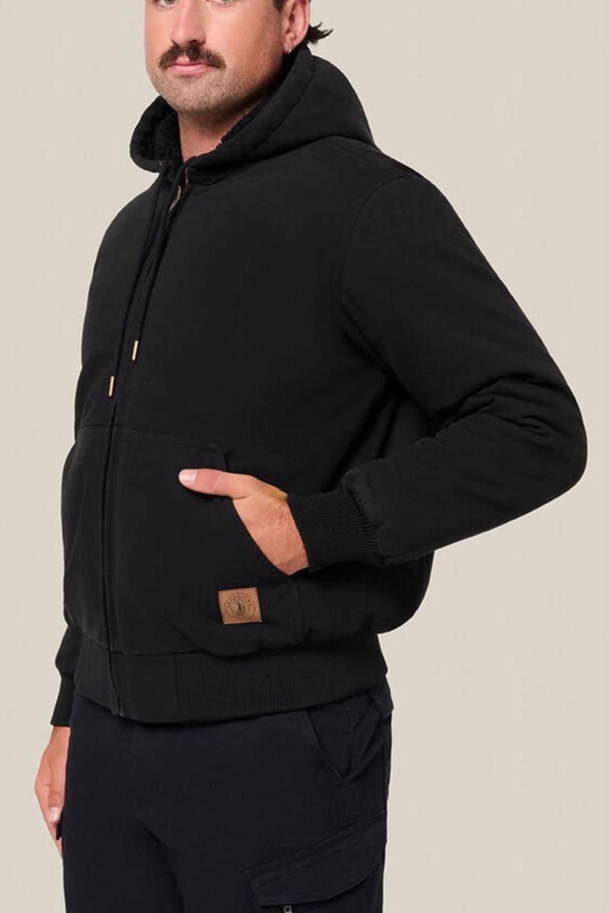 A person in a black Hard Yakka Canvas Bomber Jacket with front pockets and black pants stands against a beige background, one hand in a pocket and the other relaxed by their side.