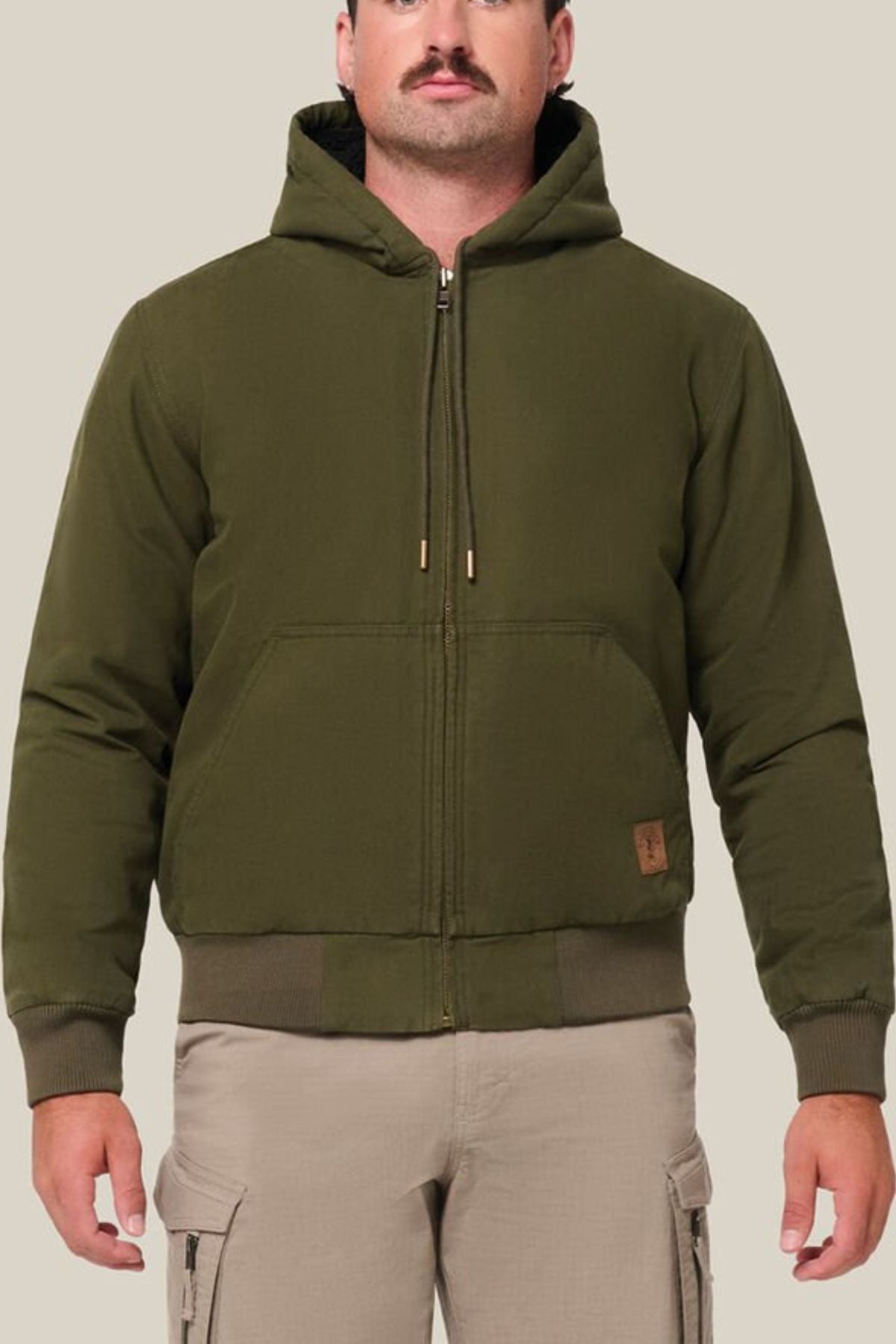 A man with a mustache wears a Hard Yakka Canvas Bomber Jacket—an olive green hooded zip-up with front pockets—and light beige cargo pants, standing against a plain light beige background.