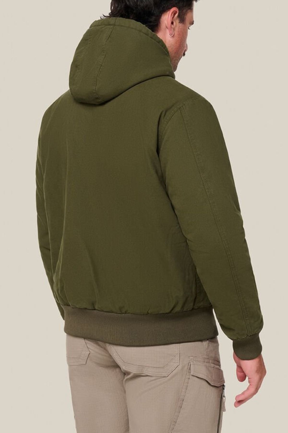 A man faces away, wearing the Hard Yakka Canvas Bomber Jacket in olive green and light beige cargo pants against a plain light background.