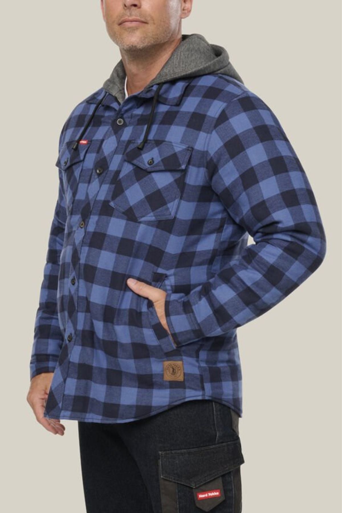A man wears a blue and black plaid Hard Yakka Quilted Flannelette Shacket with a gray hood, black pants, and his left hand in his pocket, standing against a plain beige background.