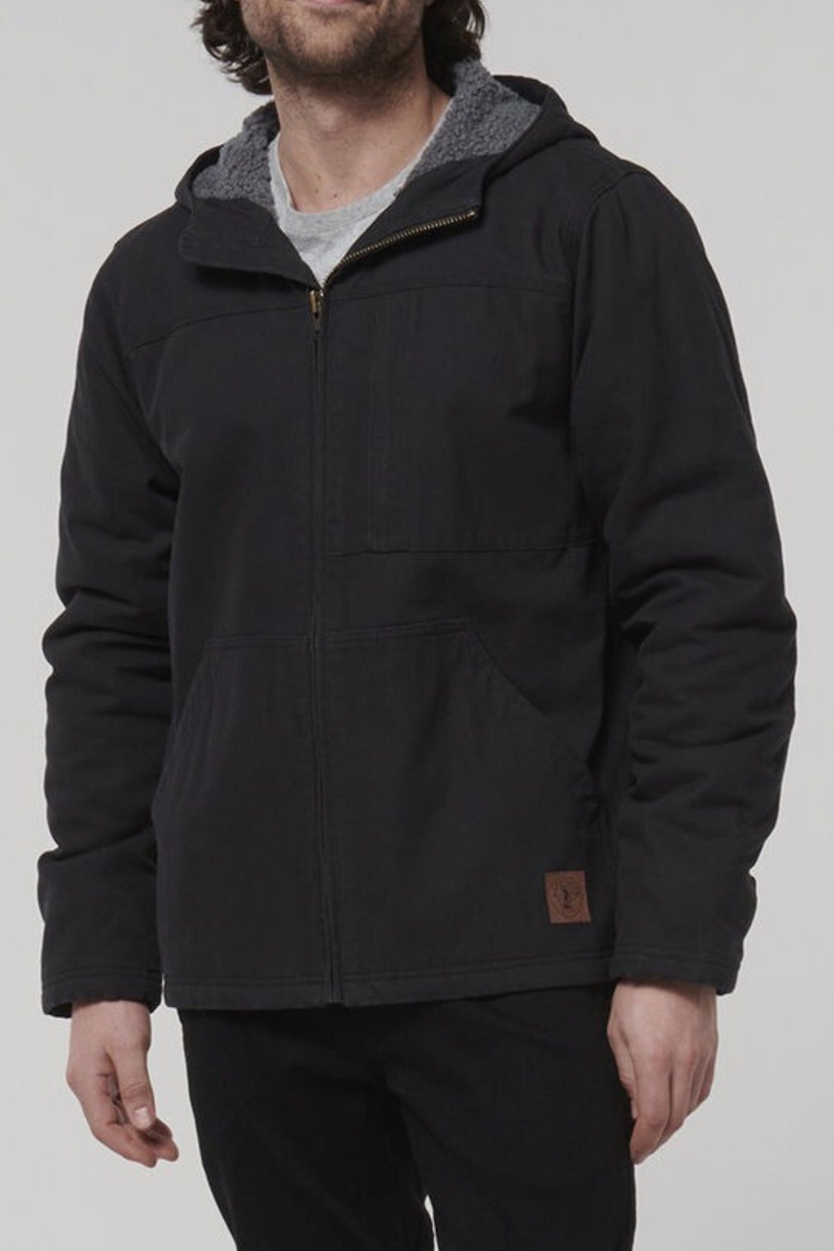 A person wears a Hard Yakka Heritage Canvas Hooded Jacket in black with sherpa lining, heavy-duty zip, and side pockets. Beneath is a white shirt paired with black pants. Their head is slightly tilted, and their hands rest at their sides.