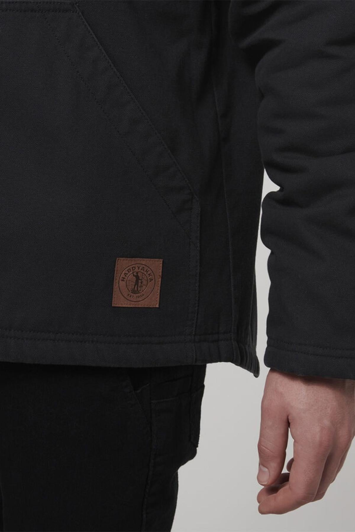 A close-up shows a person in a Hard Yakka Heritage Canvas Hooded Jacket with a circular patch, sherpa fleece lining, and heavy-duty zip. They stand with one hand by their side, also wearing dark pants.