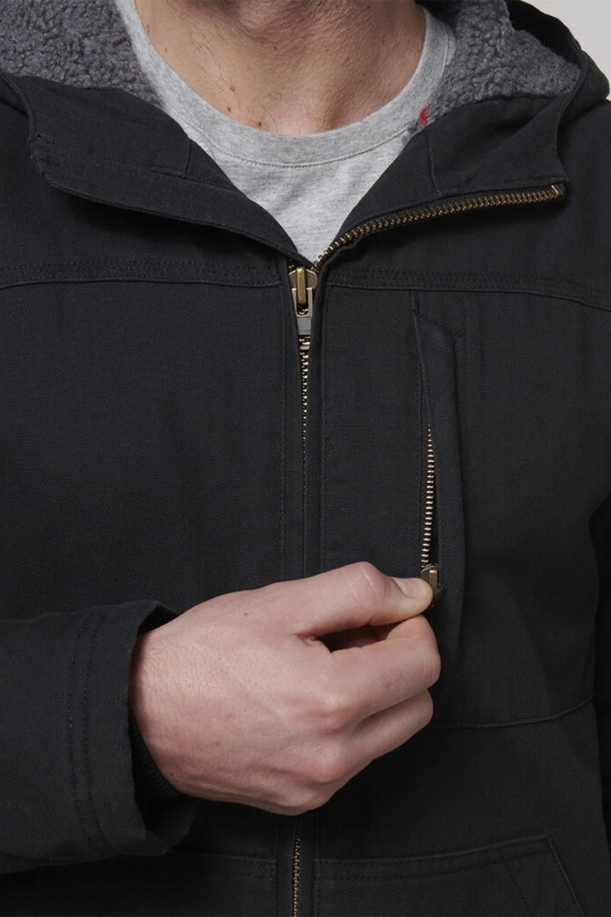 A person wearing the Hard Yakka Heritage Canvas Hooded Jacket with a gray shirt is zipping the heavy-duty brass chest zipper. The jacket's sherpa fleece lining inside the hood ensures warmth and comfort.
