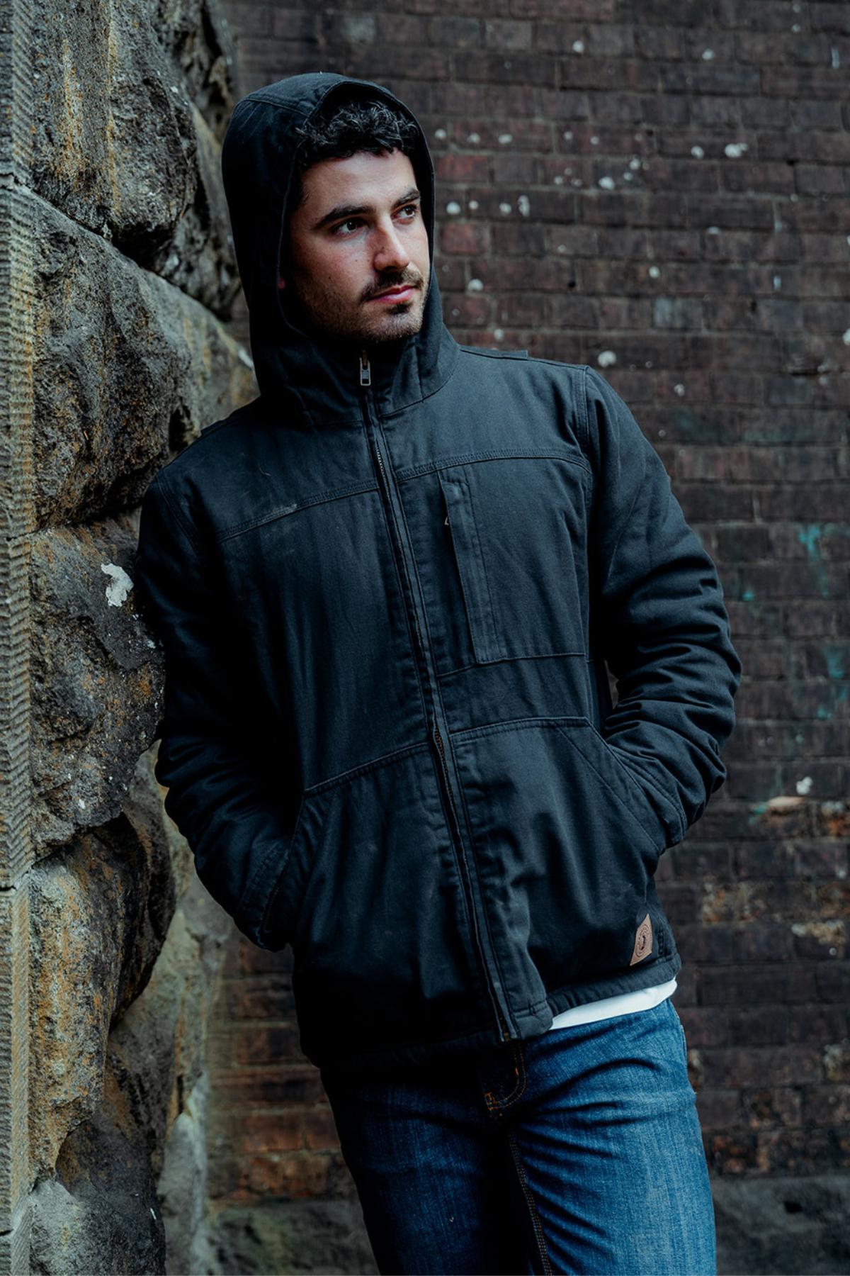 A person wears a Hard Yakka Heritage Canvas Hooded Jacket with sherpa fleece lining, standing against a stone wall. The textured brick backdrop complements the cozy warmth and style of their heavy-duty zip-up attire.