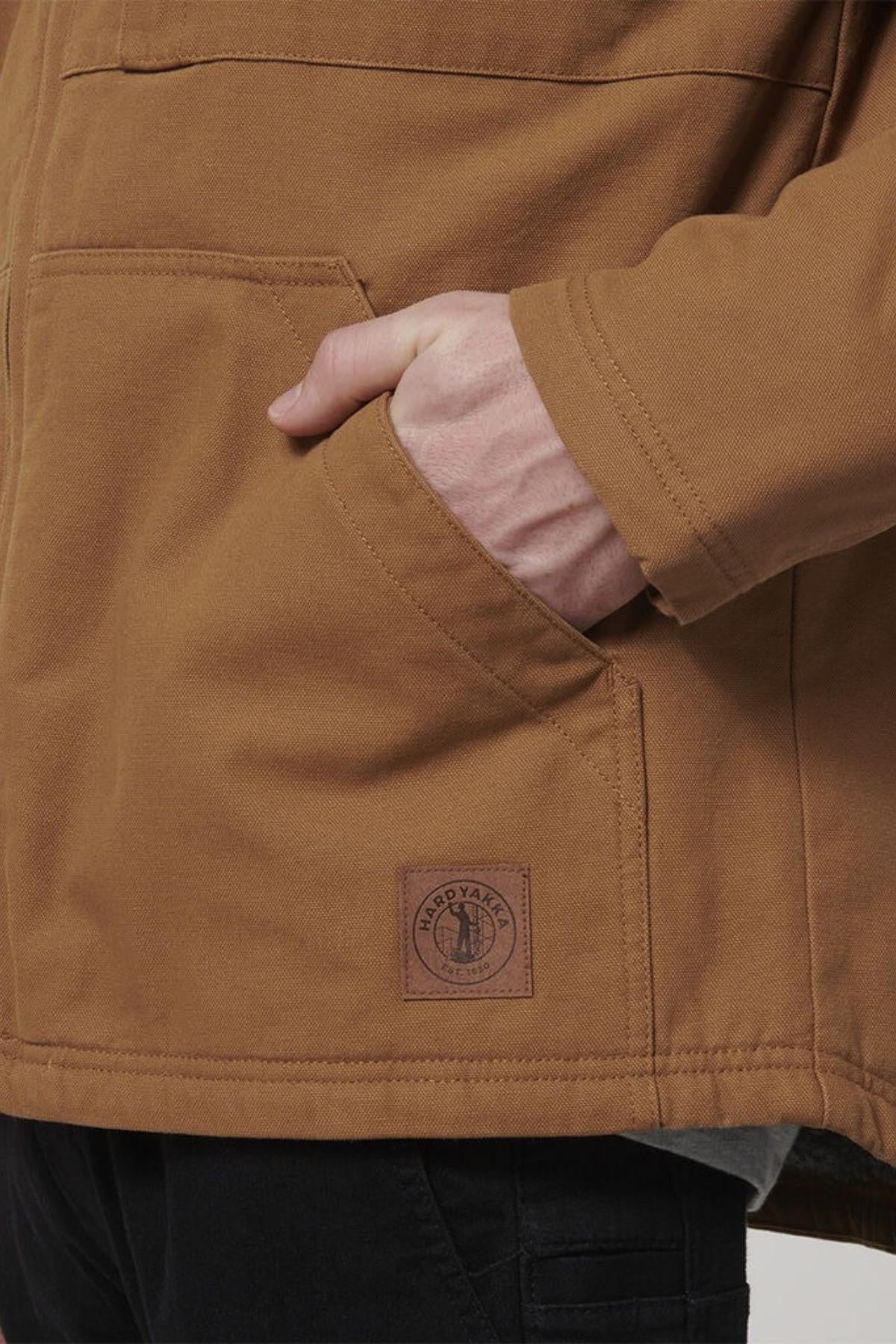 A person wears the Hard Yakka Heritage Canvas Hooded Jacket, featuring durable stitching and a visible patch near the pocket, with their right hand in the pocket, styled with black pants.