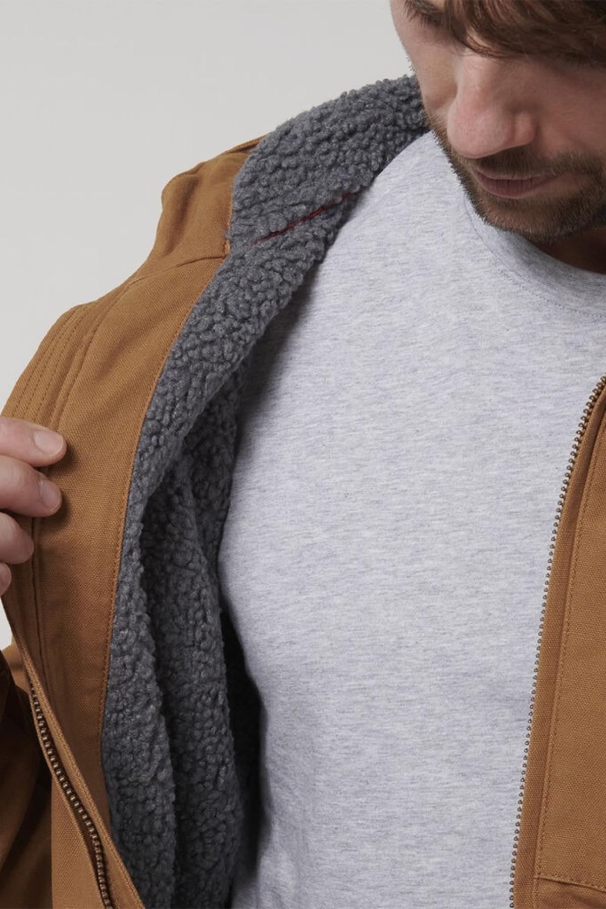 A man opens a brown Hard Yakka Heritage Canvas Hooded Jacket, showing its soft, dark gray sherpa fleece lining. The light gray shirt and durable stitching highlight the jacket’s comfort and quality. His face is partially visible as he looks down.