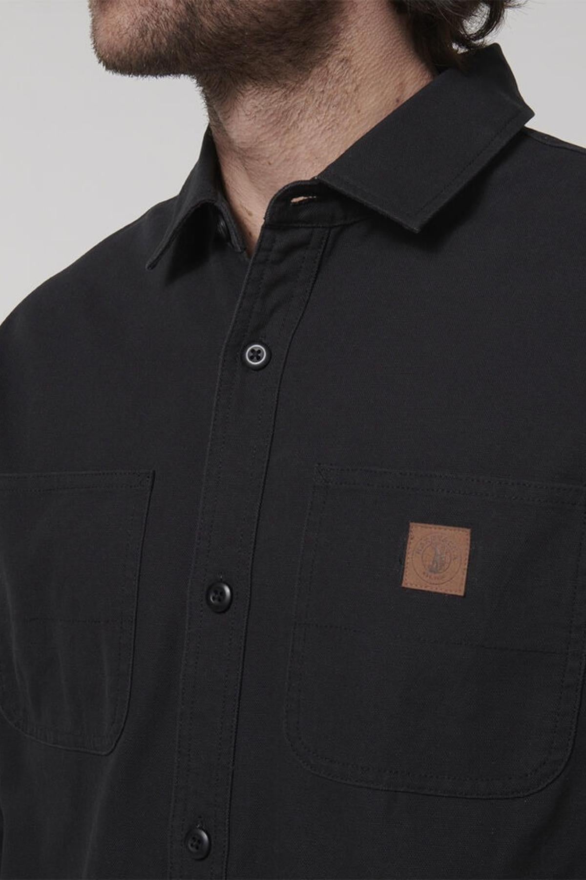 A person is seen shoulders-down wearing a durable black Hard Yakka Heritage Canvas Shirt with double chest pockets, each sporting a small brown logo patch on the left pocket, set against a plain and light-colored background.