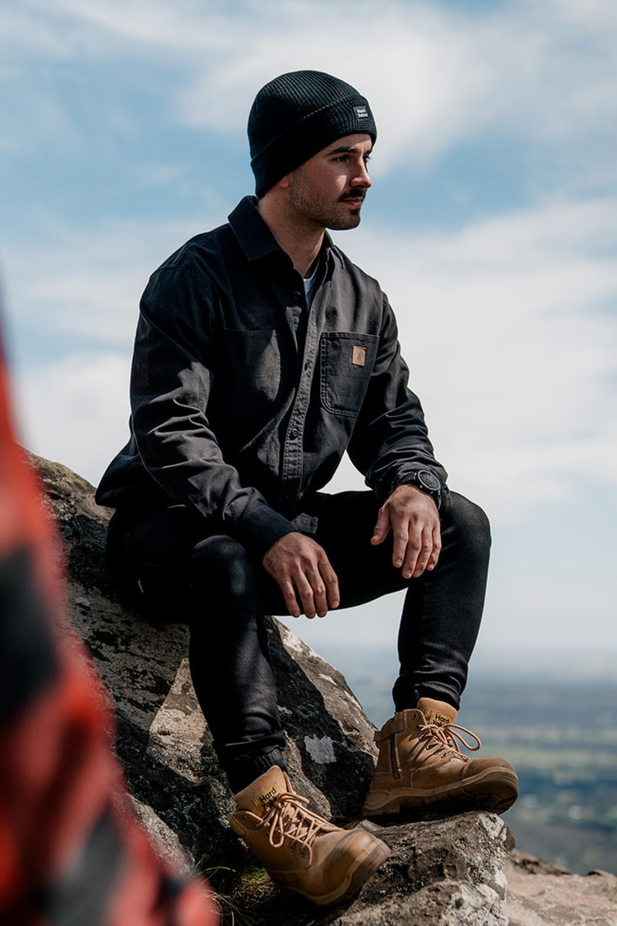 A man in a black jacket, beanie, and tan boots sits on a rock with a scenic backdrop. His Hard Yakka Heritage Canvas Shirt with double chest pockets complements the clear sky dotted with clouds.