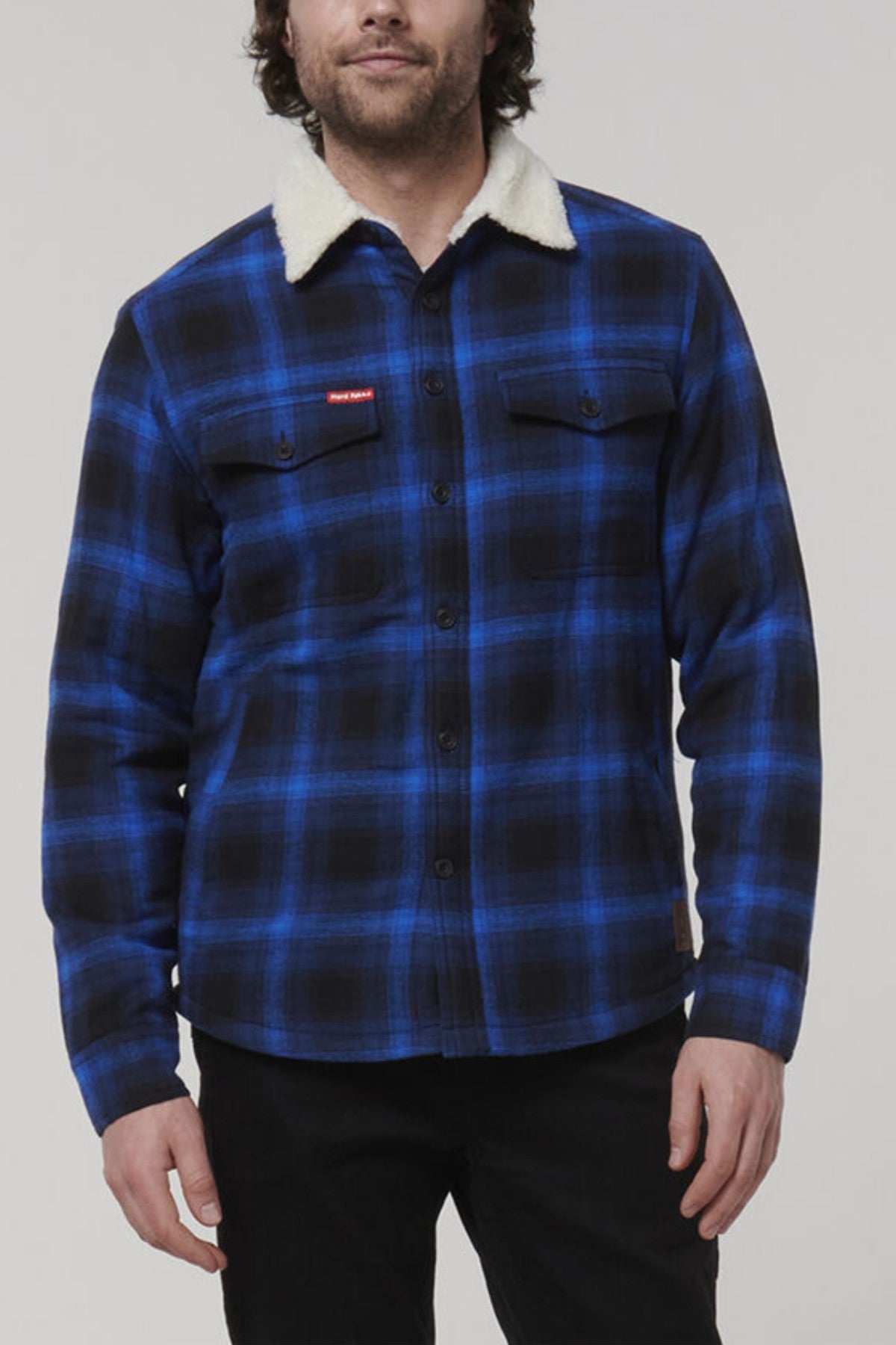 A man stands against a plain background wearing a Hard Yakka Heritage Trucker Sherpa Jacket in blue and black plaid. This regular fit jacket boasts a cozy sherpa collar, button-up closure, and practical chest pockets, merging functionality with style.