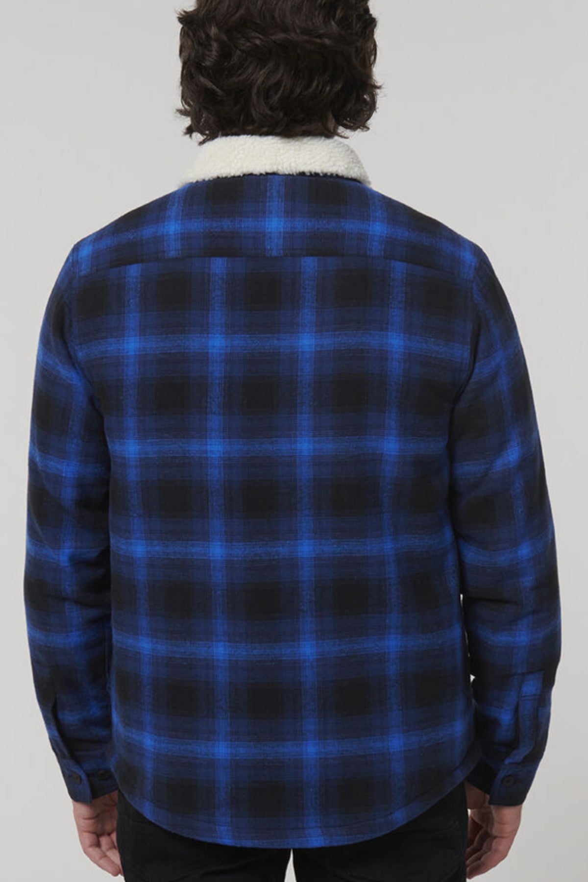 The image features a person from the back wearing a blue and black plaid Hard Yakka Heritage Trucker Sherpa Jacket with a white sherpa collar, dark curly hair, and standing against a light gray background.