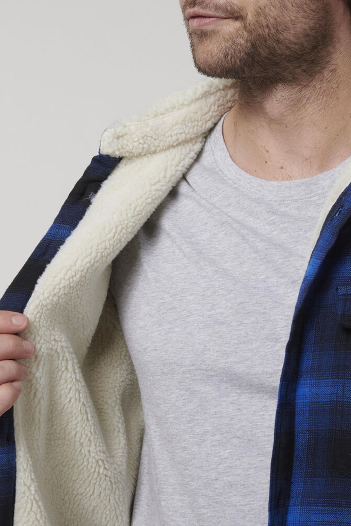 The photo shows a person in the Hard Yakka Heritage Trucker Sherpa Jacket, featuring a regular fit with a blue and black plaid pattern and a cozy sherpa collar. It's partially open to reveal a gray T-shirt, adding rugged style and framing part of the person’s face and upper torso.