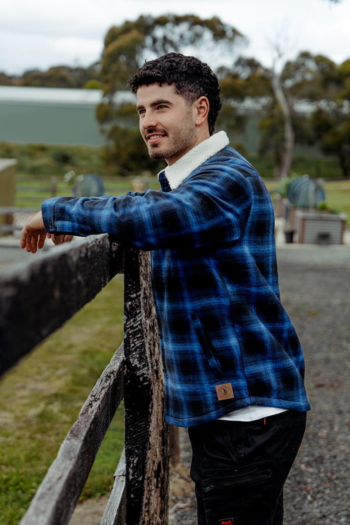 A man in a blue plaid jacket, the Hard Yakka Heritage Trucker Sherpa, leans on a wooden fence gazing into the distance. The scene features grassy areas and trees with blurred structures in the cloudy backdrop, while the jacket's sherpa collar adds cozy style.