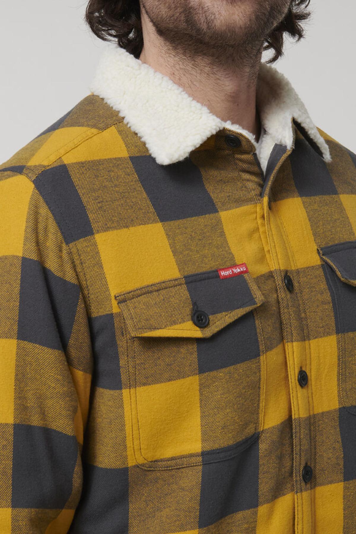 A person wears the Hard Yakka Heritage Trucker Sherpa Jacket—a yellow and dark gray plaid flannel with a white sherpa collar, black buttons, red chest pocket tag, and 100% cotton. Only the lower half of their face is visible.