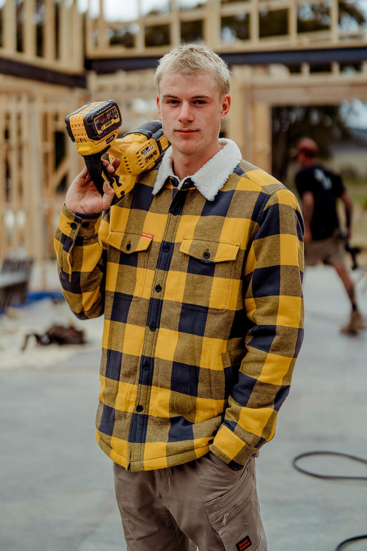 A young man wears a Hard Yakka Heritage Trucker Sherpa Jacket and holds a power drill over his shoulder at a building site with wooden framing, while another worker is visible in the background.
