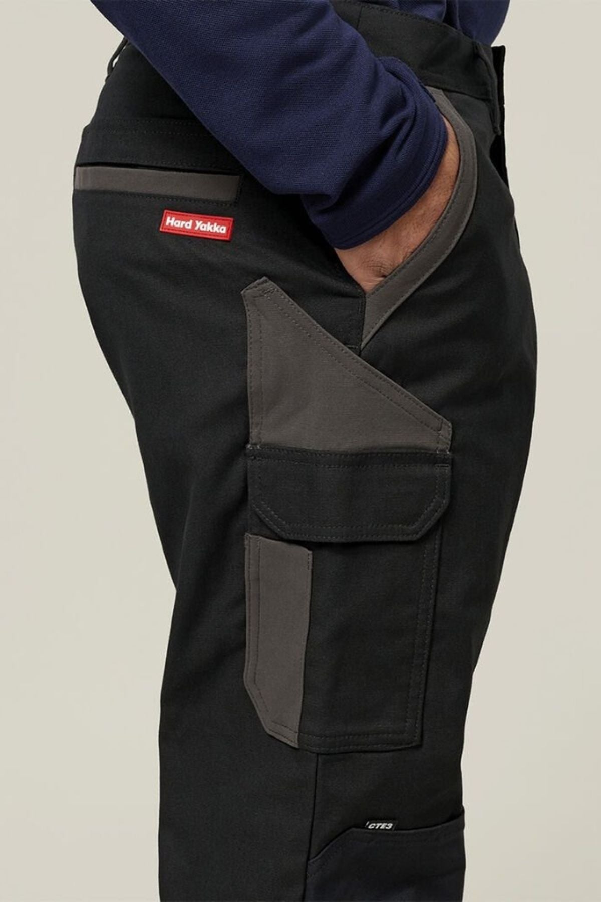 A person wearing Hard Yakka Legends Slim Stretch Work Pant in black and dark gray, featuring reinforced stretch fabric, a large side pocket, and a red Hard Yakka logo tag; shown waist down with one hand in their pocket.