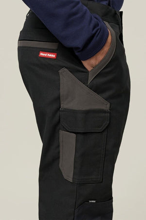 A person wearing Hard Yakka Legends Slim Stretch Work Pant in black and dark gray, featuring reinforced stretch fabric, a large side pocket, and a red Hard Yakka logo tag; shown waist down with one hand in their pocket.