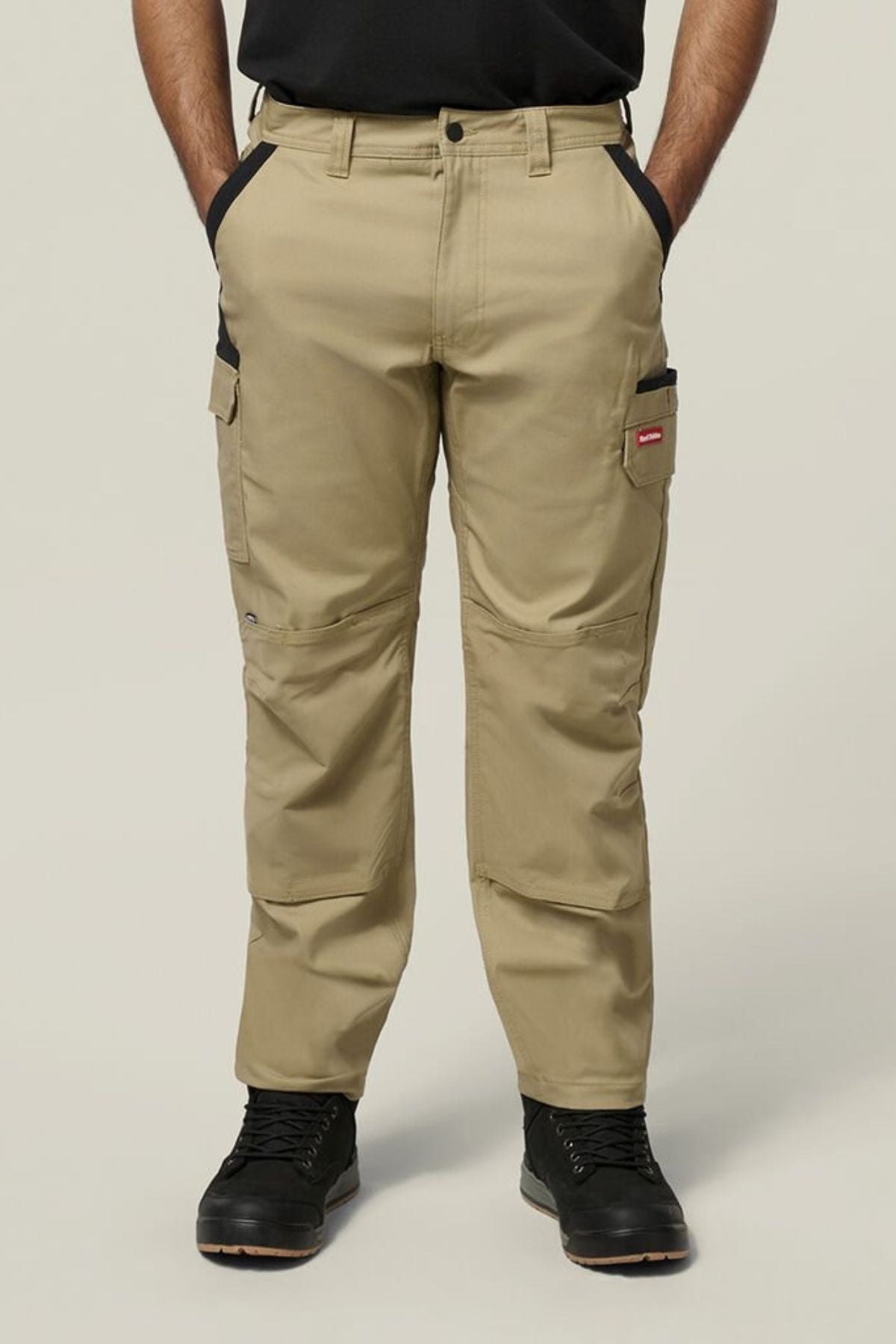 A person wearing tan Hard Yakka Legends Slim Stretch Work Pant with multiple utility and cargo pockets, paired with black boots, stands facing forward with hands in the front pockets against a plain light-colored background.