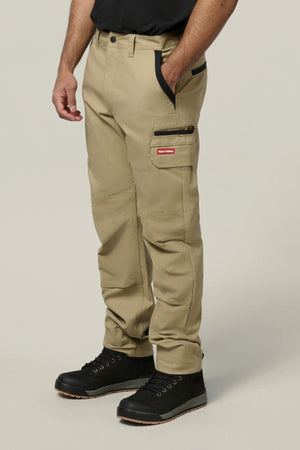 A person wears Hard Yakka Legends Slim Stretch Work Pant with black accents and utility pockets, featuring a red and white label on khaki fabric, against a plain light background, paired with black shoes.