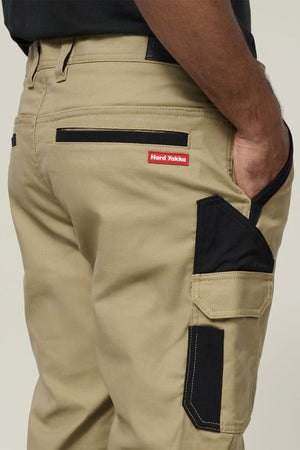 A close-up of a person wearing Hard Yakka Legends Slim Stretch Work Pant in khaki, featuring black accents, utility pockets, and a red and white label above the back pocket; their right hand is in the side pocket.