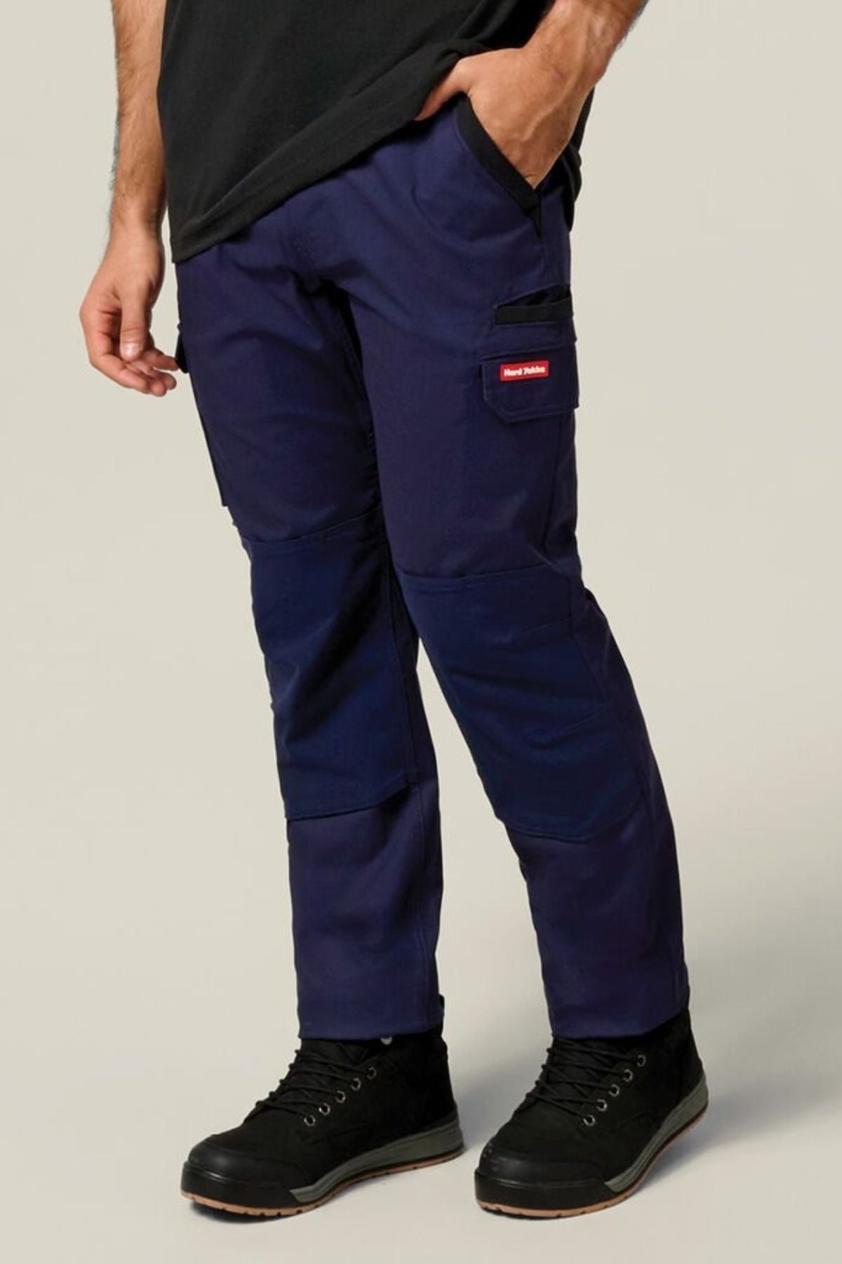 A person wearing Hard Yakka Legends Slim Stretch Work Pant in dark blue, featuring reinforced knees and utility pockets, along with black shoes and a black shirt with one hand in the pocket, stands against a plain light background.