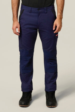 A person wearing Hard Yakka Legends Slim Stretch Work Pant in navy with utility and spacious side pockets, reinforced knees, plus black boots and a black shirt stands against a plain light background.