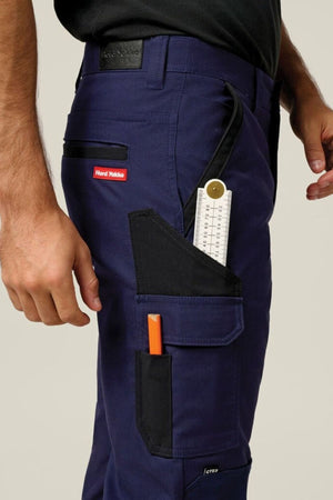 A person wears the Hard Yakka Legends Slim Stretch Work Pant, storing a ruler and orange pen in the black and red thigh utility pockets. Their arm rests at their side, paired with a black shirt.