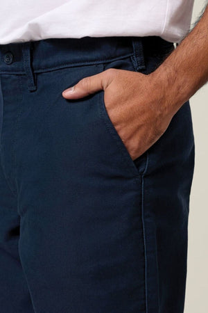 A person in a white shirt and dark blue Hard Yakka Moleskin Plain Front Heavy Duty Work Pant stands with one hand casually in their pocket; only their torso and upper legs are visible.