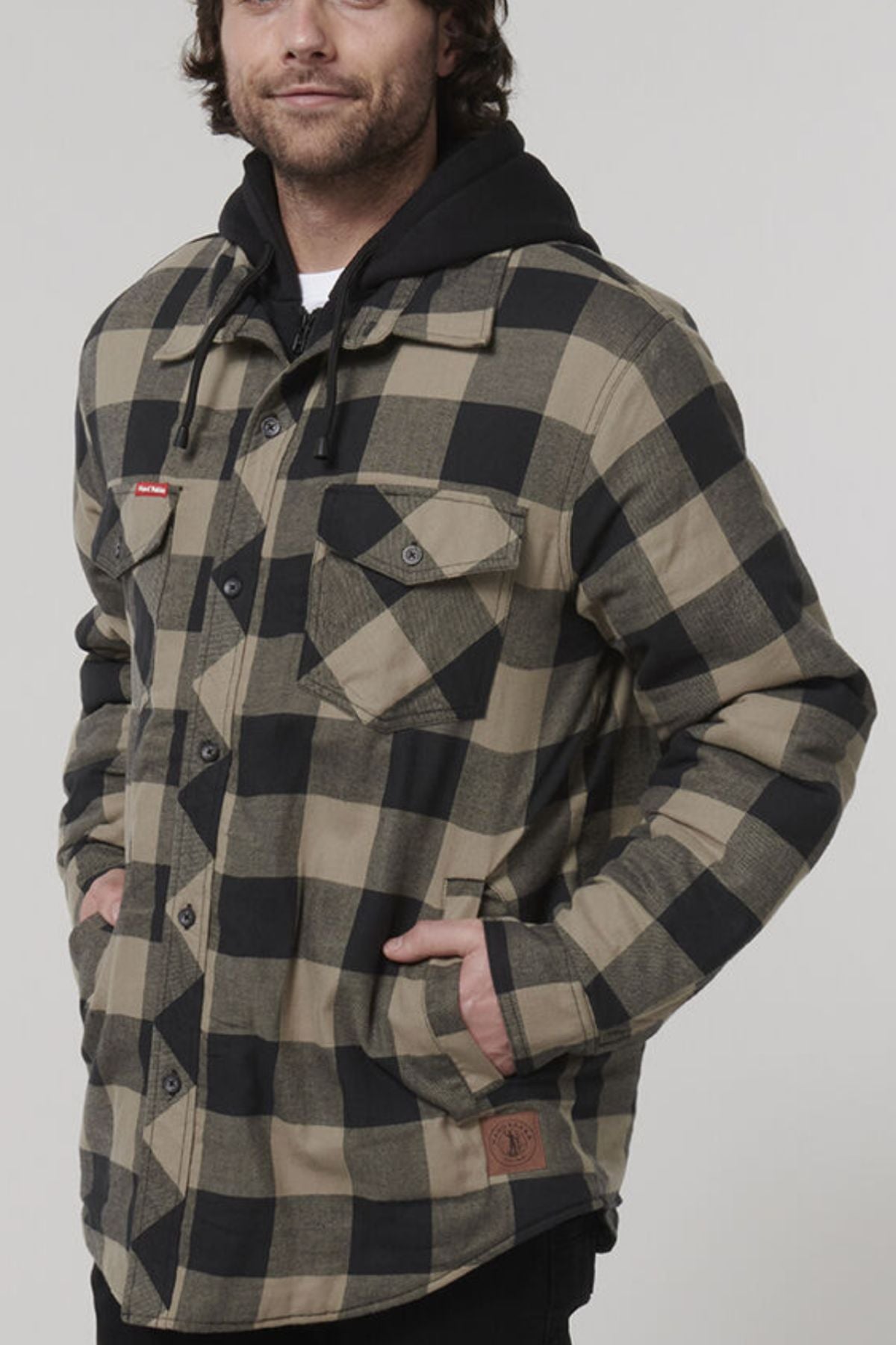 A person sporting the Hard Yakka Quilted Flannelette Shacket, a durable black and beige checkered jacket, layered over a black hoodie, stands with hands in pockets. This shacket includes two chest pockets fastened with buttons. The backdrop is plain and light-colored. The individual has shoulder-length hair and wears casual attire.