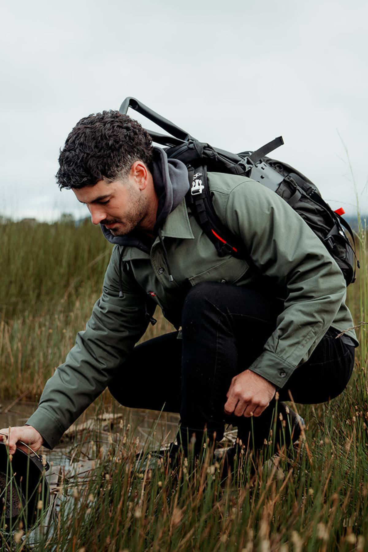 A man with curly dark hair, wearing a Hard Yakka Quilted Solid Shacket made from REPREVE® material, black pants, and a backpack, crouches in a grassy wetland area, reaching down with his left hand towards the water. The overcast background features more grasses and a distant landscape.