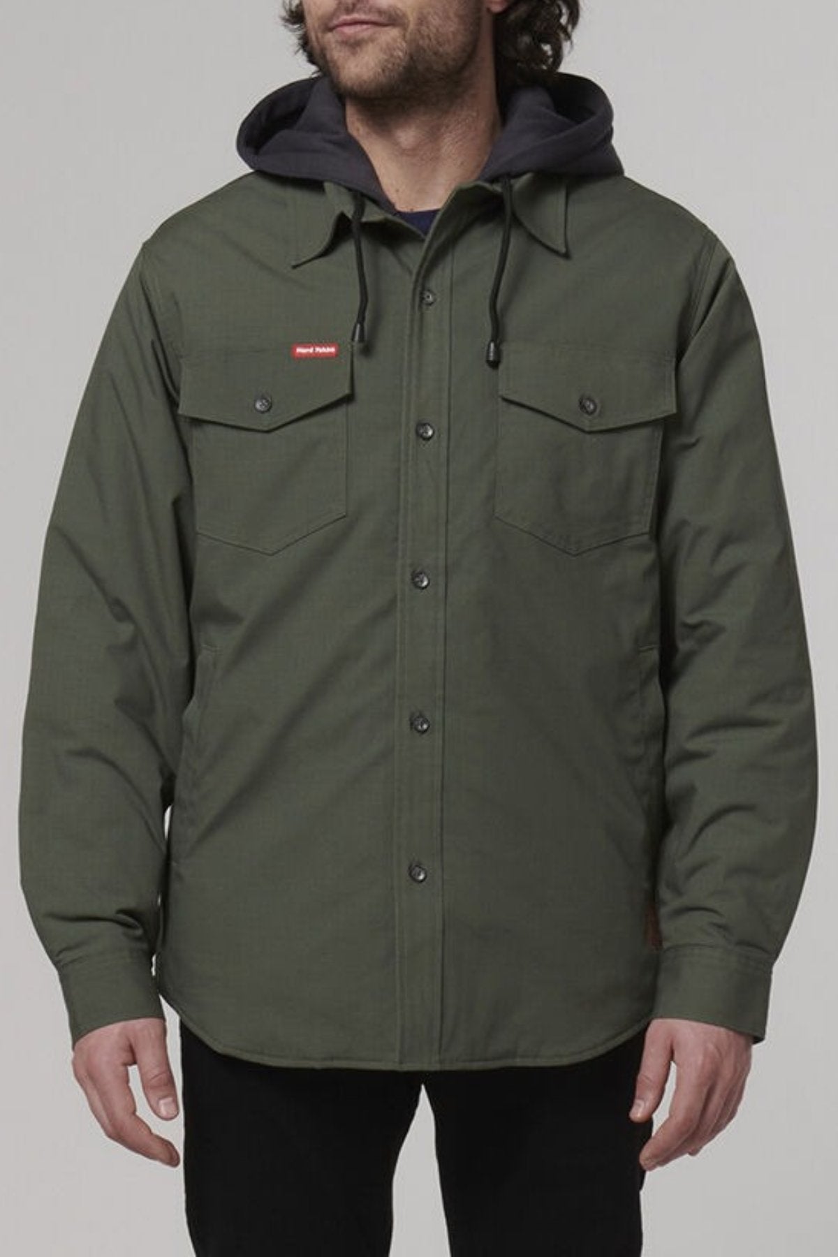 A person is wearing a Hard Yakka Quilted Solid Shacket in green, featuring two chest pockets and a small red logo on the left chest. The jacket is made from durable ripstop fabric, and they're layering it over a black hoodie. The background is plain and light grey.