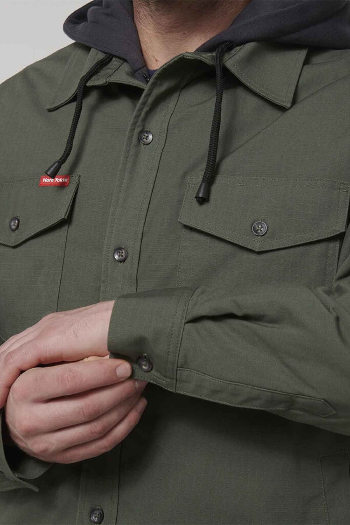 A person wearing the Hard Yakka Quilted Solid Shacket in olive-green, with black drawstrings visible from the hooded sweatshirt underneath. The shacket, made from durable ripstop fabric, features buttoned chest pockets and the person is fastening the cuff of their left sleeve. A small red label is sewn on one of the pockets.